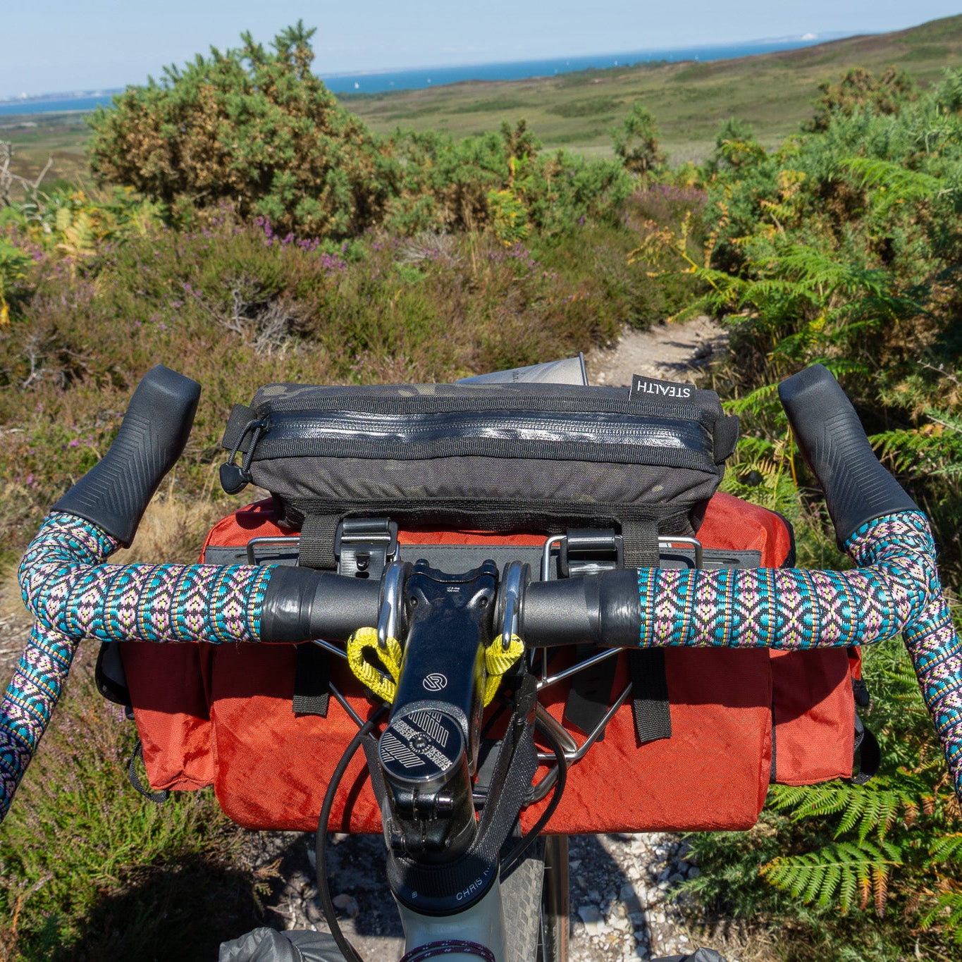 Bicycle handlebars with colorful grips and a red bag on a trail with greenery and a clear sky.